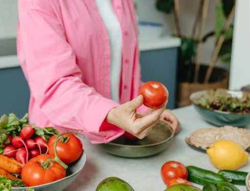 Person in pink overshirt holding a fresh tomato with various organic vegetables on a countertop.